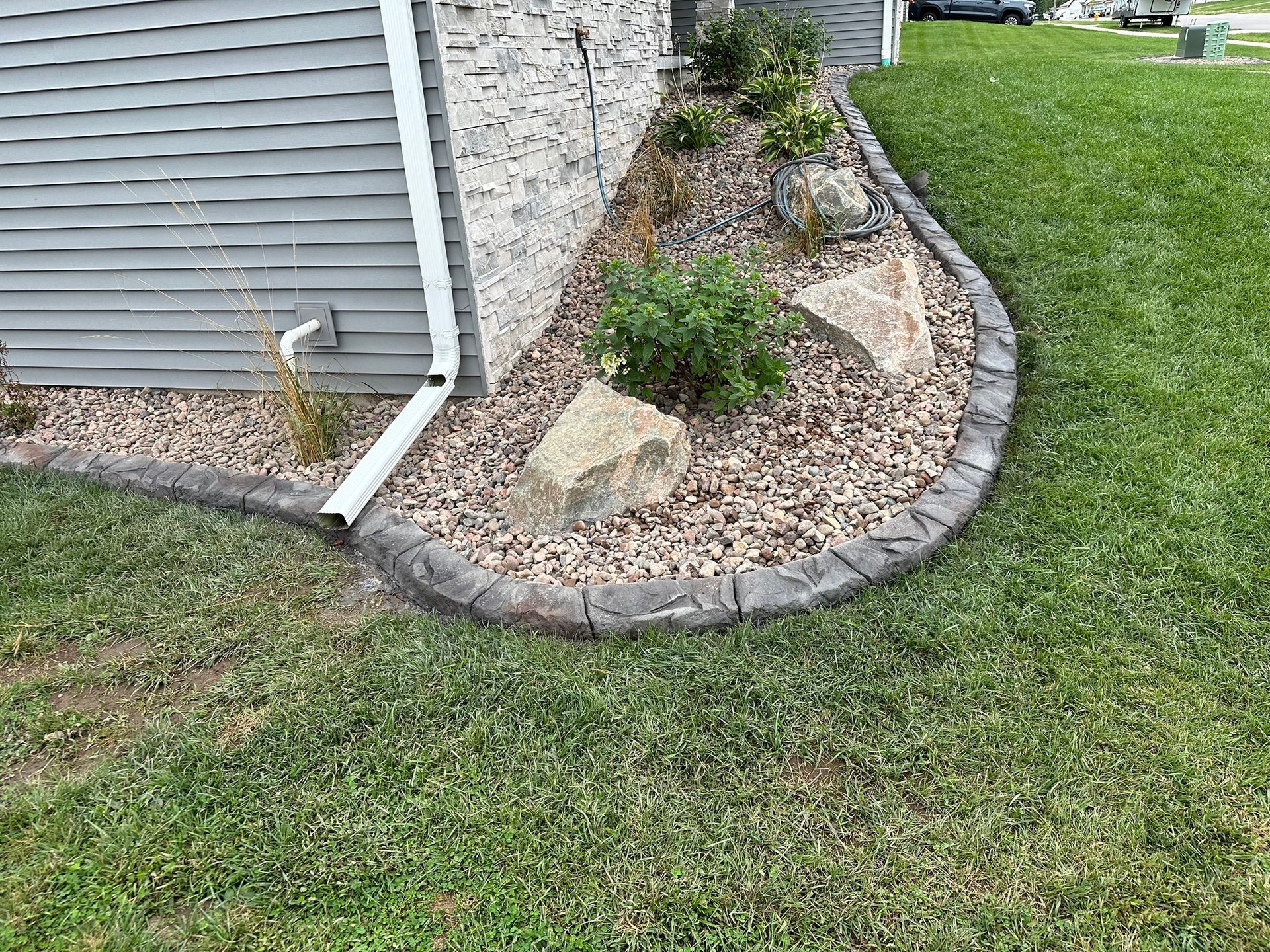 Curved gray concrete border separates a stone-filled planting bed from the lawn beside a home