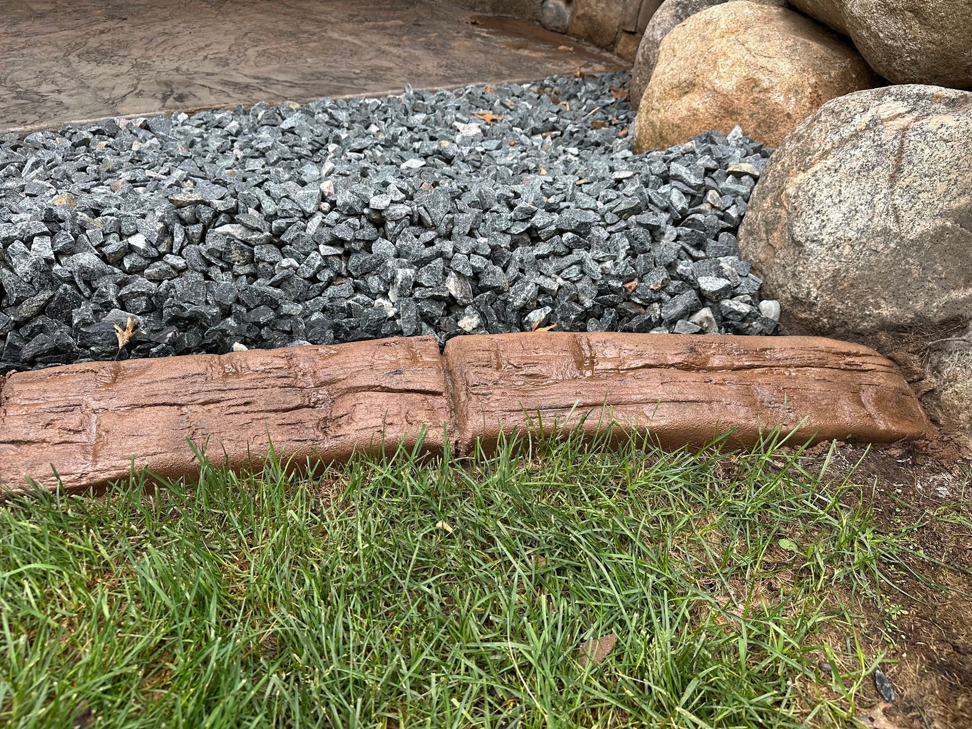 Warm brown wood-grain concrete curbing alongside a gray stone landscape bed